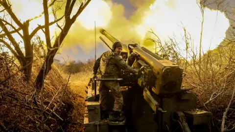 EPA A Ukrainian soldier turns his face away from the blast as a giant cannon he is operating fires. He is surrounded by trees.