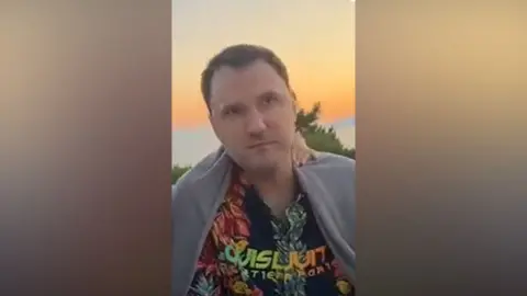 Other Vitaly Nikolayevich Kovalev wears a vibrant Hawaiian style shirt in front an orange and yellow sunset sky.