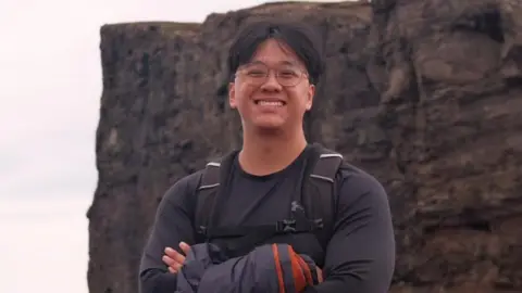 Shyan Lim Shyan Lim smiling for a picture with his arms crossed during a hike near a cliff