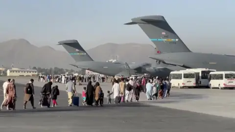 BBC People carrying luggage walk across an airport tarmac towards large US military transport aircraft during an evacuation