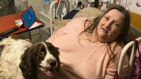 Comfort Companions CIC Yvonne Stride, a woman wearing a pink jumper is in bed with her dog on her lap and her i-pad with the Comfort Companions app on her bedside table.