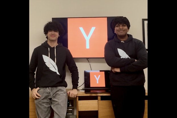Ishaan Gangwani (l) and Aayam Bansal, two teenagers wearing black hoodies with large white feather logos on front. They're standing in front of a desk with a laptop and a wall-mounted TV. The laptop and TV screens show a large orange box with a white Y in the middle.