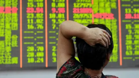 Reuters An investor looks at an electronic board showing stock information with numbers highlighted in red and lime green against a black background. The investor has her back to the camera and has her left hand resting on her head.
