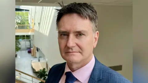 NHSBT Head of organ transplantation at NHS Blood and Transplant, Anthony Clarkson. He is stood upstairs overlooking a building atrium. He is wearing a blue suit, pink shirt and blue tie. He is looking directly at the camera.