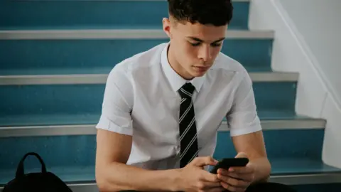 Getty Images an adolescent boy in a short-sleeved shirt and a tie sitting on stairs, looking at a phone