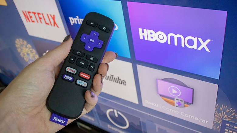 A hand holding the Roku remote next to a TV
