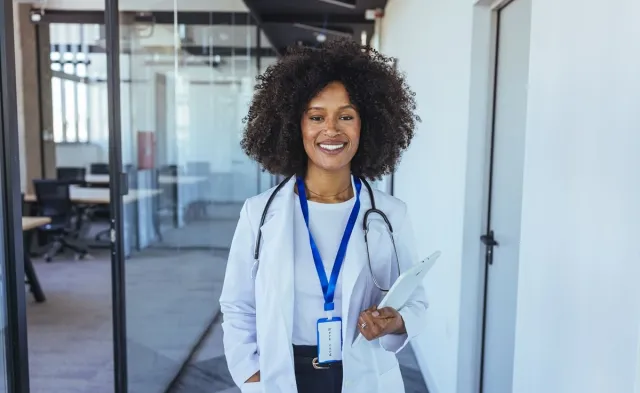 Smiling nurse practitioner in a white coat with a stethoscope holds a tablet in a modern medical office.