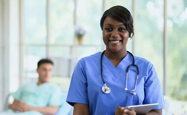 Smiling nurse in blue scrubs with a stethoscope and tablet standing in a bright hospital room, with a patient resting in a bed blurred in the background.