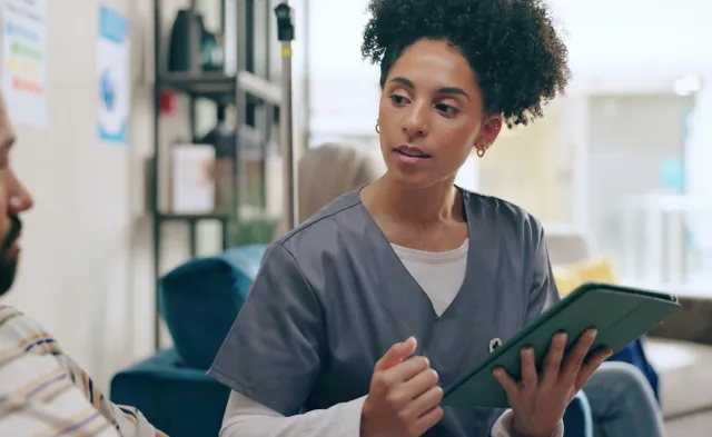 Registered nurse who graduated from an MSN Direct Entry program in scrubs discusses information on a tablet with a seated man in a home setting.