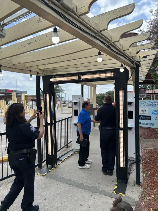 The pergola has lights hanging from the rafters. Two men are standing underneath black metal scaffolding that has additional lighting sources. They are standing near one of the grey metal kiosks. A female Border Patrol officer is a few feet behind the men, and she is taking a cell phone picture of them. Although we can’t see her face well, it appears she might be smiling.