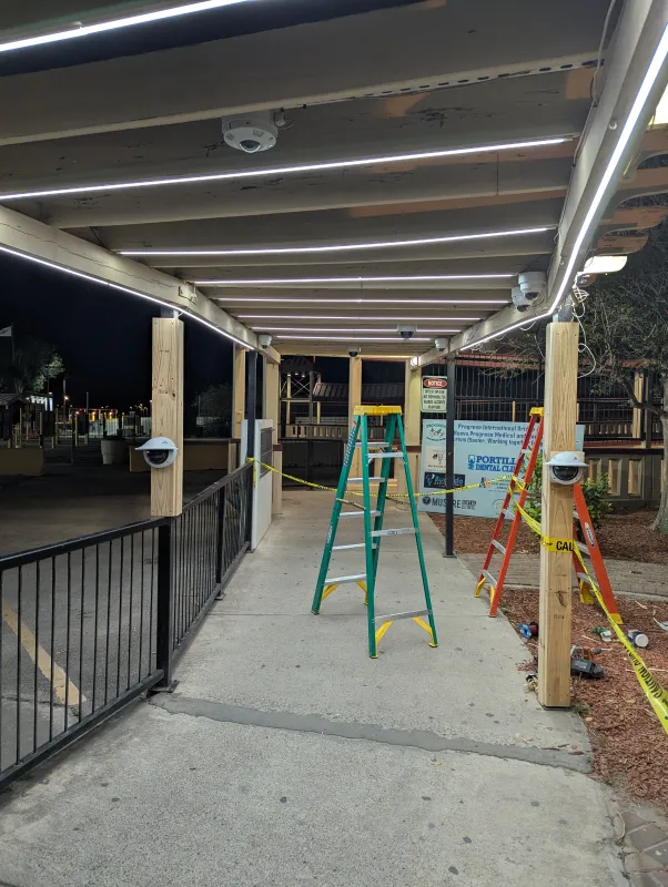A green and orange ladder, and yellow “Caution” tape wound around the poles and across the walkway. Looking down the weathered wooden pergola pedestrian walkway, there are cameras on the support poles and in the rafters. There are lighting strips affixed to the rafters as well as other lighting sources.